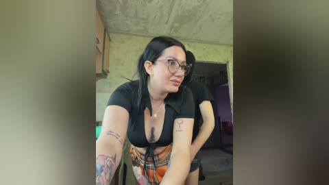 A video of a plus-sized woman with fair skin, black hair, glasses, and tattoos, wearing a low-cut black top and plaid skirt, leaning forward in a dimly lit room with green walls and exposed ceiling.