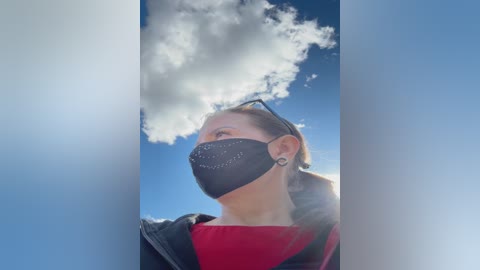 A video of a young woman with light skin and blonde hair, wearing a black face mask and black jacket over a red shirt, gazing upward at a bright blue sky with fluffy clouds.