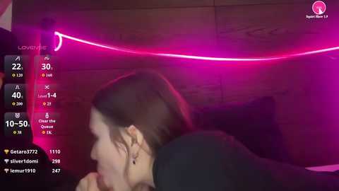 Video of a woman with light skin and brown hair lying on her stomach, wearing a black top, in a dimly lit room with neon pink lights and a digital interface showing live stream stats.