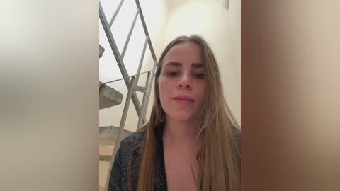 Video of a young Caucasian woman with long, straight blonde hair, wearing a patterned cardigan, standing in a modern, minimalistic room with a metallic staircase and white walls.