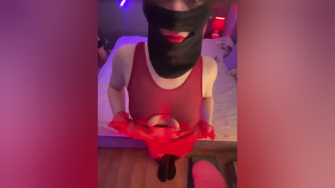A video of a person in a red dress, black mask, and gloves, standing in a dimly-lit room with a bed and red light.