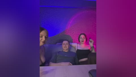 Video of a man and woman in a dimly lit, purple-lit room; the man, with short hair, sits smiling, while the woman, with short hair, stands behind him.