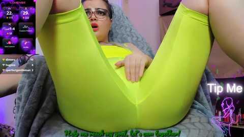 A video of a young woman with glasses and bright yellow leggings, lying on her back with legs spread, revealing her crotch, in a cozy, dimly lit room.
