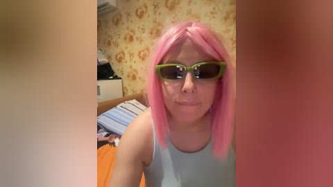 A video of a person with pink hair, wearing green-tinted 3D glasses and a light blue tank top, in a room with floral wallpaper and a bed with orange and blue striped bedding.