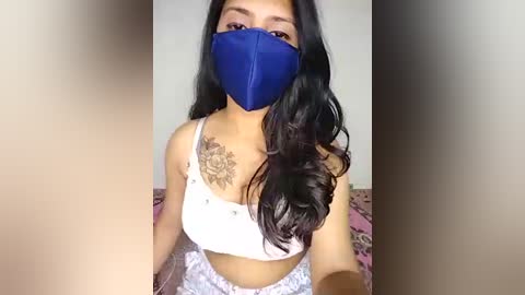 A video of a young woman with long, wavy black hair, wearing a blue mask, white bra, and patterned skirt. She has a floral tattoo on her shoulder. Background shows a pink-patterned bedspread.