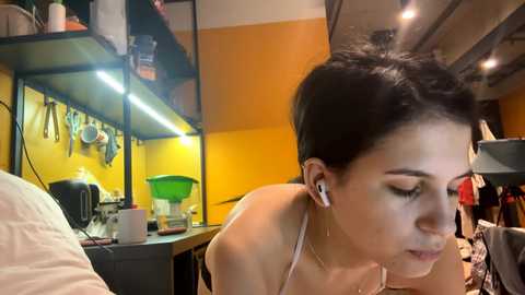 Video of a young woman with short black hair, wearing white headphones, leaning on a kitchen counter in a modern, well-lit room with yellow walls and exposed pipes.