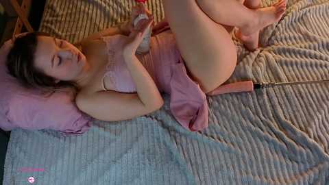 Video of a young, light-skinned woman with brown hair, wearing a pink lace bra, lying on a bed with a pink blanket, holding a pink vibrator.