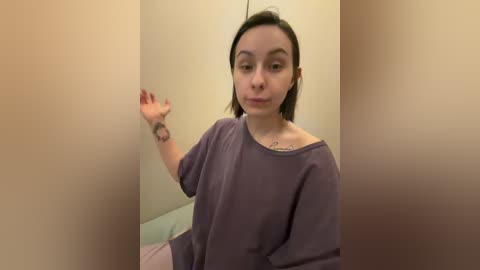 A video of a young woman with short brown hair, wearing a loose, purple T-shirt, standing in a beige-walled room. She gestures with her right hand, revealing a tattoo on her wrist.
